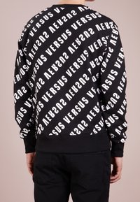 Felpa nera con testo "VERSUS" bianco ripetuto. Maniche e orlo a costine, e vestibilità rilassata. Tessuto morbido con un audace design grafico.