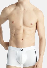 Weiße Baumwoll-Boxershorts mit einem schwarzen elastischen Bund, der das Adidas-Logo trägt. Enganliegende Passform, glatte Textur und seitliche schwarze Akzente.