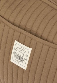 Brūna dūraiņu auduma ar vertikālām šuvēm, uz kuras ir krēmkrāsas logotipa uzlīme ar mājas ilustrāciju un tekstu "GAI + LIVA DENMARK".