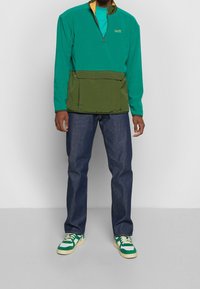 Suéter bloqueado en colores verde y turquesa con media cremallera, gran bolsillo frontal, combinado con jeans azul oscuro y zapatillas con acentos verdes.