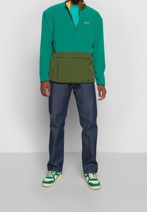 Grøn og turkis farveblokket pullover med en halvlynlås, stor frontlomme, parret med mørkeblå jeans og sneakers med grønne accenter.