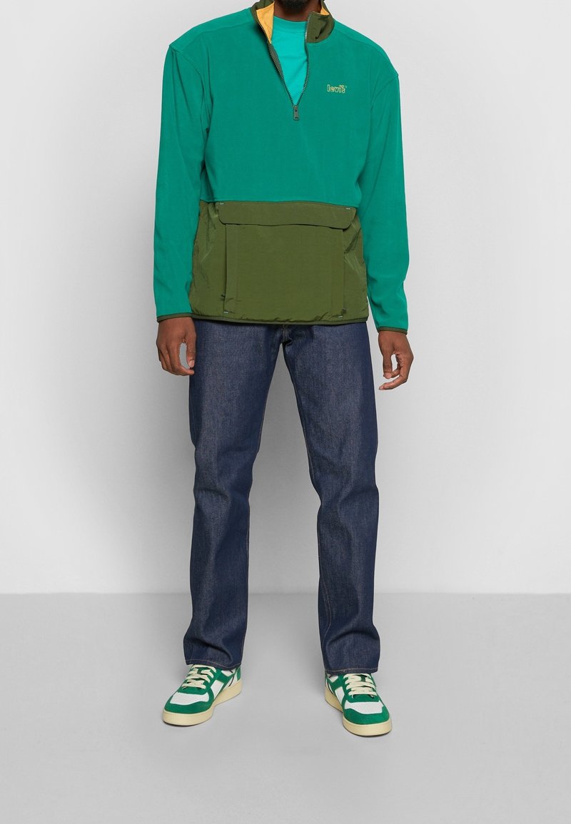 Suéter bloqueado en colores verde y turquesa con media cremallera, gran bolsillo frontal, combinado con jeans azul oscuro y zapatillas con acentos verdes.