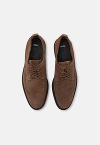 Scarpe da dress in suede marrone con classici dettagli brogue, punte rotonde e suole in gomma nera piatta. I lacci sono marroni, con il marchio BOSS all'interno.
