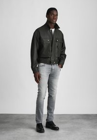 Μαύρο δερμάτινο μπουφάν με ψηλό γιακά, μπροστινό φερμουάρ και πολλαπλές τσέπες, συνδυασμένο με ανοιχτό γκρίζα τζιν και μαύρα παπούτσια.