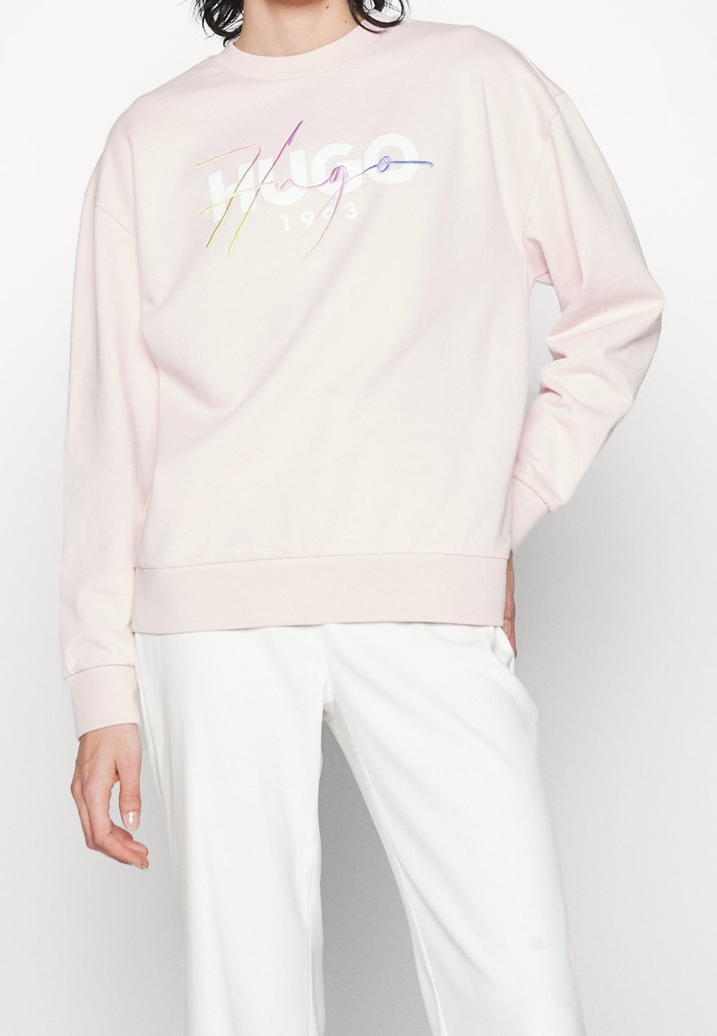 Personne portant un sweat-shirt rose pâle avec un logo multicolore "Hugo 1993" et un pantalon blanc, debout avec une main dans la poche.