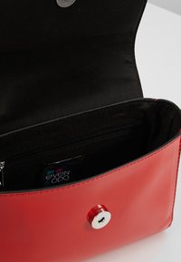 Bolso de cuero rojo con acabado brillante, que cuenta con un interior de tela negra, un bolsillo con cremallera y un detalle de cierre con botón circular.