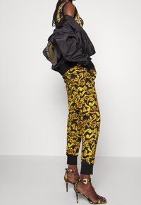 Svart bomberjacka över en gul mönstrad outfit med svarta accenter, kombinerad med gula och svarta sandaletter med hög klack.