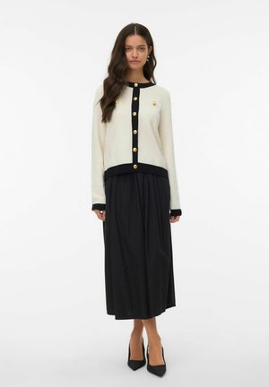 Cream cardigan with black trim and gold buttons paired with a black flowy skirt, featuring a relaxed fit and textured fabric.
