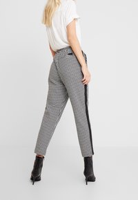 Kostkované kalhoty v černobílé barvě se střihnutím rovné nohavice, zdobené kontrastními bočními pruhy a vysokým pasem. V kombinaci s černými kotníkovými botami.