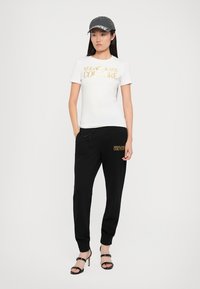 Camiseta blanca con texto dorado "Versace Jeans Couture", combinada con pantalones de chándal negros y relajados que presentan un logo a juego. Gorra gris y tacones negros.