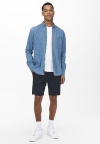 Blaue Denim-Button-Up-Hemd über einem weißen T-Shirt, kombiniert mit dunklen Shorts und weißen Turnschuhen. Das Modell steht mit den Händen in den Taschen.