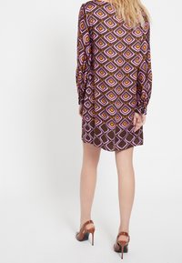 En person med långt hår bär en mönstrad brun klänning med lila och orange diamanter, i kombination med bruna högklackade skor.