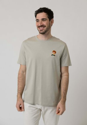 Mann lächelt, trägt ein hellgraues T-Shirt mit kleinem buntem Cartoon-Charakter auf der linken Brust, kombiniert mit weißer Hose vor einfachem Hintergrund.