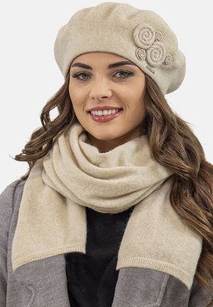 Béret beige avec un design de rosette et écharpe beige en tissu doux ; les deux présentent une texture lisse. Le modèle porte un manteau gris en dessous.