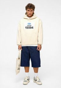 Beige hoodie met een grafische print, denim shorts, witte sokken en sneakers. Een natuurlijke tote bag hangt aan de linkerhand.