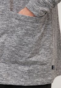 Mano parzialmente dentro la tasca frontale di un maglione grigio a maglia testurizzata.