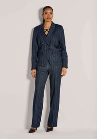 Vrouw die tegen een lichte muur staat, gekleed in een marineblauwe gestreepte dubbele blazer, bijpassende broek en zwarte puntige hakken.