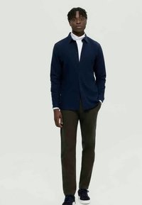 Marineblauwe button-up shirt, witte coltrui, donkergroene broek en donkere sneakers. Model staat tegen een lichtgrijze achtergrond.