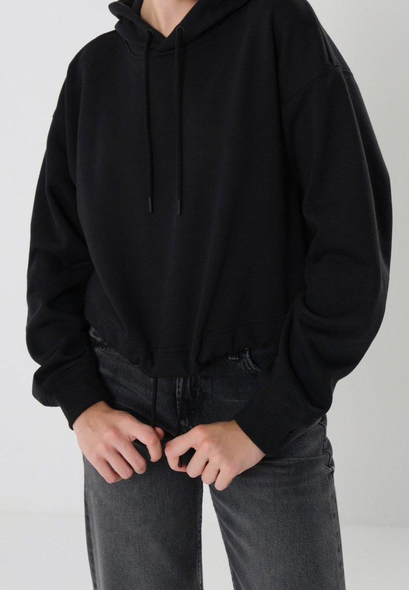 Sudadera corta negra hecha de una tela suave, con capucha ajustable, puños acanalados y dobladillo elástico. Combinada con jeans gris oscuro.