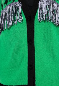 Neulecardigan kirkkaan vihreänä, mustalla nappilista, johon on lisätty mustavalkoista fringepitimä reunojen ympärille.