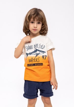 Kurzärmliges T-Shirt in Creme und Orange mit einem Hai-Motiv und Text, kombiniert mit navyblauen Shorts. Glatte Baumwollstruktur.
