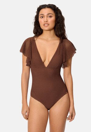 Mujer con un bañador de una pieza marrón con textura, escote en V profundo y mangas con volantes, de pie frente a un fondo liso y claro.