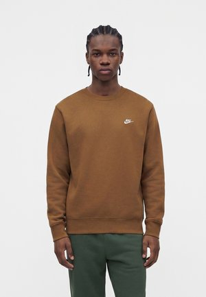 Homme aux cheveux tressés portant un sweat-shirt Nike marron et un pantalon vert, debout devant un fond clair uni.