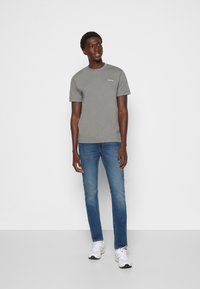 Camiseta de algodón gris con mangas cortas y cuello redondo, con un logo sutil. Combinada con jeans rectos azules y zapatillas blancas.