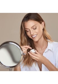 Crema per la cura della pelle in un piccolo barattolo bianco tenuto da una persona con lunghi capelli ondulati. La persona indossa una camicia bianca e sta usando uno specchio.