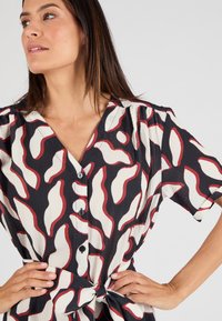 Camicia con bottoni di base nera, caratterizzata da un motivo astratto a onde rosse e crema. Ha maniche corte e un dettaglio con vita annodata.