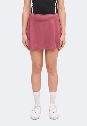 Persona con top negro, falda de tenis malva, calcetines blancos de caña alta y zapatillas deportivas blancas, de pie con los brazos relajados a los lados.