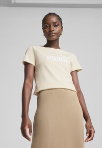 Beige bomulds t-shirt med hvid "PUMA" logo, parret med en tan nederdel. Korte ærmer og en afslappet pasform. Glat tekstur, minimalistisk design.