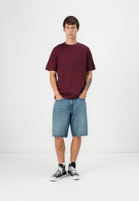 Bordeauxrotes T-Shirt mit kurzen Ärmeln, blaue Jeansshorts und schwarze High-Top-Sneaker. Einfaches Design mit lockerer Passform und sichtbaren Tätowierungen am Arm.