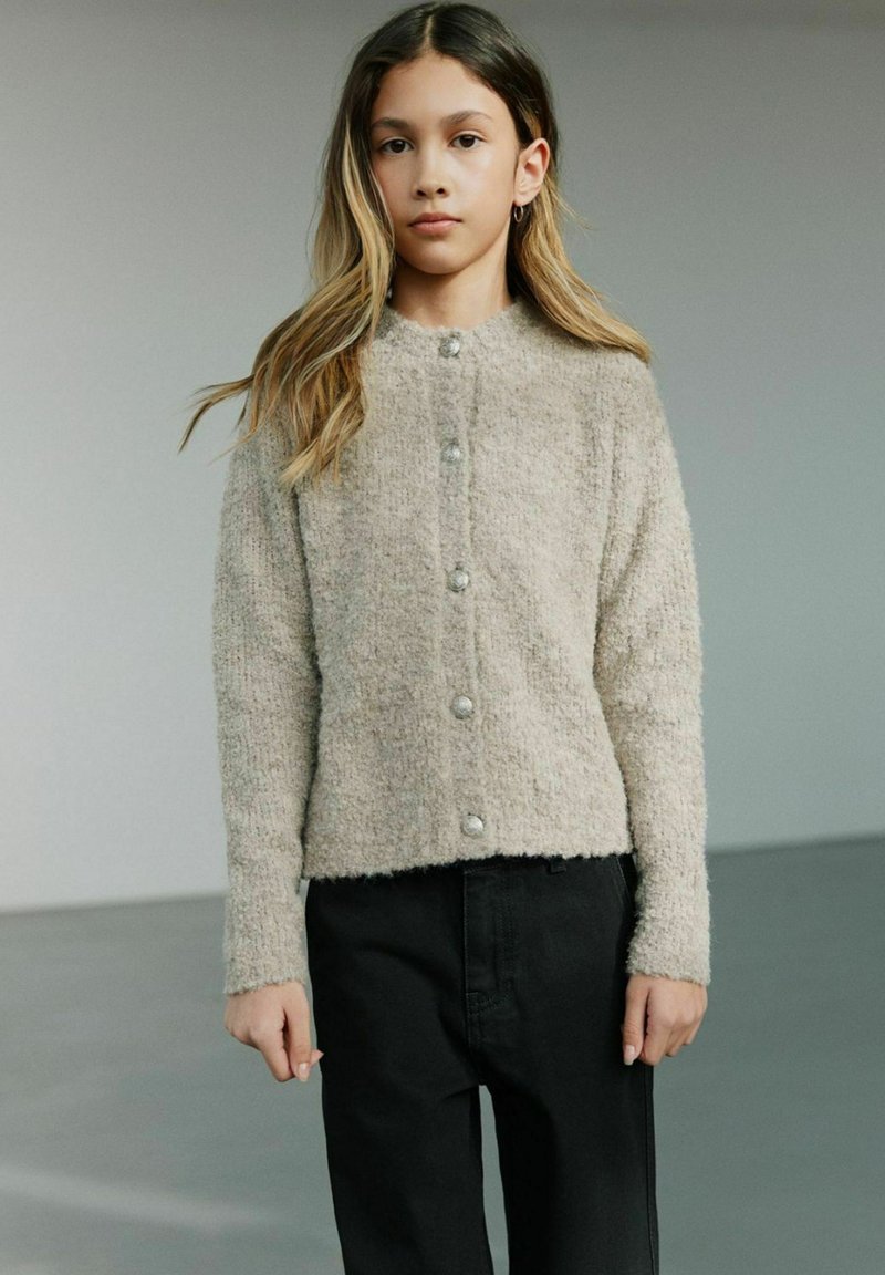 Beige fuzzy cardigan med rund halsudskæring, lange ærmer og fem store knappelukninger. Parret med mørke bukser. Glat tekstur.