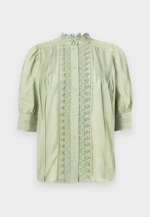 Ανοιχτοπράσινη μπλούζα με δαντέλα στο μπροστινό μέρος και στον γιακά, πιέτες στους ώμους και μισομακριά φουσκωτά μανίκια με μανσέτες από δαντέλα.