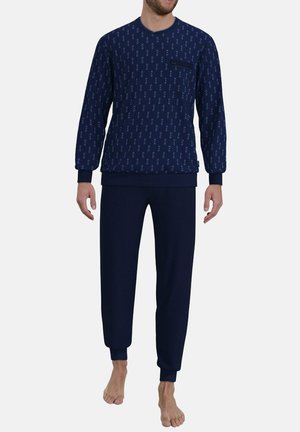 Mann trägt ein dunkelblaues Langarmoberteil mit kleinem Muster und passende einfarbige dunkelblaue Hose, steht barfuß vor einfachem Hintergrund.