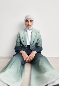 Ombre-Anzug in Grün und Marineblau, mit einem taillierten Sakko und weit geschnittenen Hosen. Getragen mit einem hellgrauen Schal und einem weißen Rollkragenoberteil.
