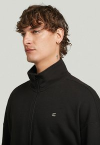 Sweatshirt noir zippé avec col haut, présentant un détail de logo subtil et un tissu lisse et texturé. Gros plan sur le col et le visage.