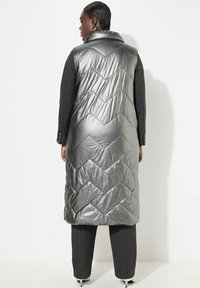 Silberne Steppweste mit Chevron-Muster, hohem Kragen und langer Silhouette, kombiniert mit dunkel gestreiften Hosen.