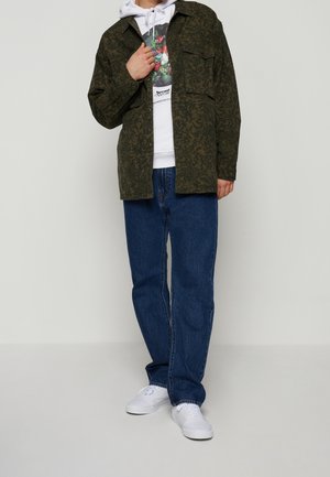 Personne portant une veste verte foncée à motifs, un sweat à capuche blanc avec un graphisme, un jean bleu et des baskets blanches, debout devant un fond gris clair.