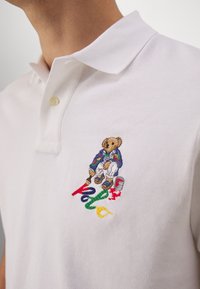 Bílá polokošile z texturované bavlny s barevnou výšivkou medvěda v modré bundě a malířskými detaily pod ní.