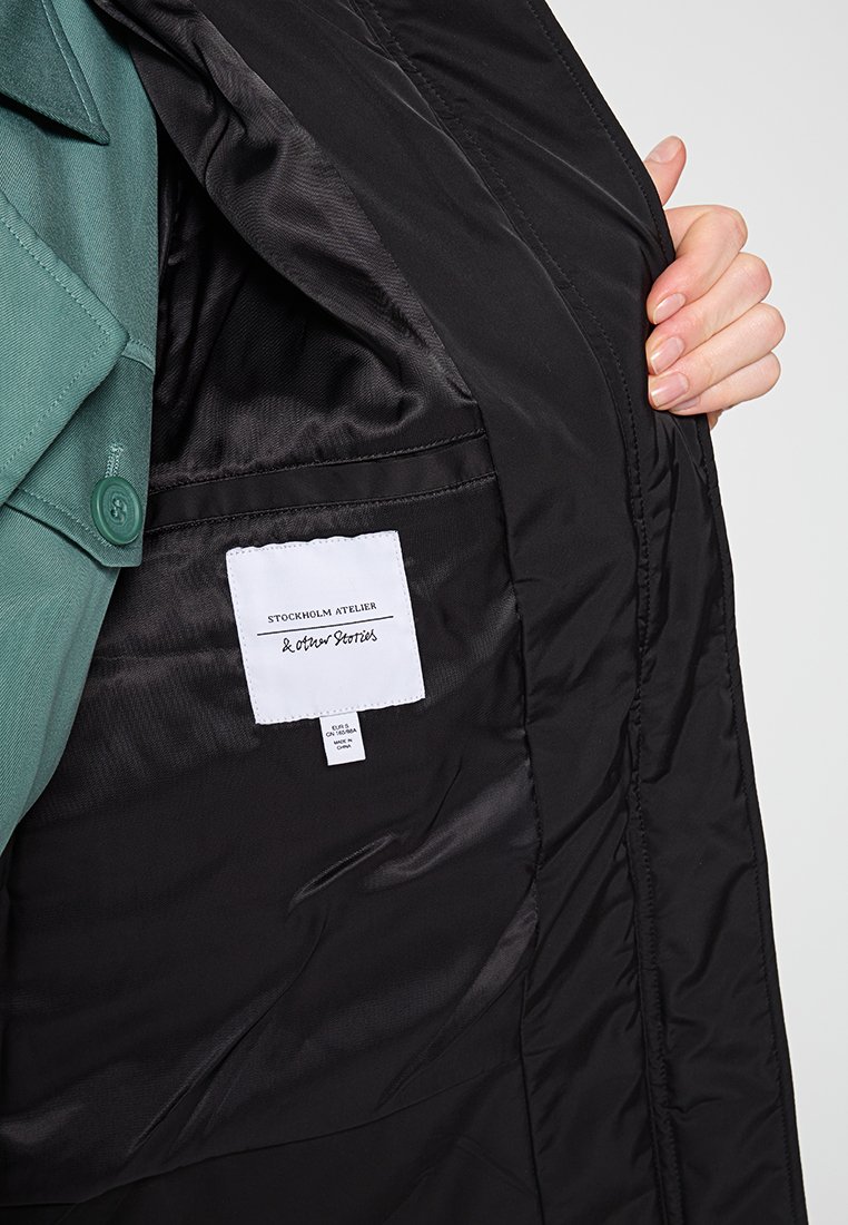 Zwarte gewatteerde jas met een gladde binnenvoering. Beschikt over een groene stoffen kraag, een label en een zijzak in de voering.