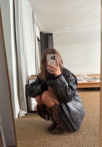 Chaqueta oversize de cuero negro con costuras blancas, superpuesta a una falda marrón con flecos. Botas marrones con flecos sobre un suelo texturizado. Reflejo en el espejo.