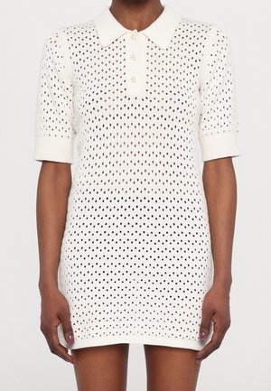 Robe en maille blanche à manches courtes avec patte de boutonnage à col et motif ajouré à pois portée par une personne à la peau foncée.