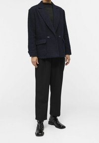 Blazer bleu marine texturé avec deux poches frontales, boutons double boutonnage, porté sur un haut noir et associé à un pantalon noir et des bottes brillantes.