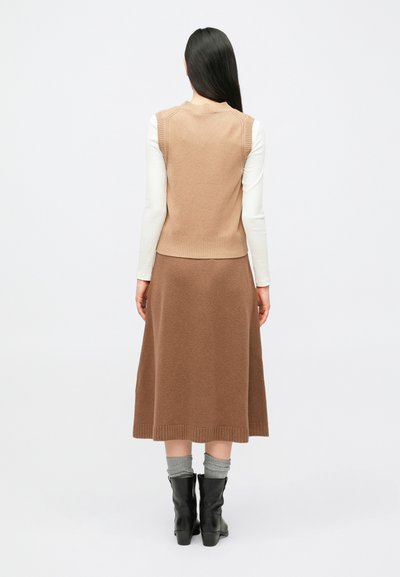 Pikkade mustade juustega naine kannab beeži varrukateta kampsunit valge pika varrukaga topi kohal, pruuni midi-seelikut, halli sokke ja musti poolsaapad.