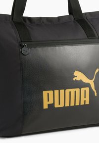 Zwarte sporttas met een getextureerd leren paneel, voorzien van een gouden 'PUMA' logo en een ritsvak. Stoffen handvatten voor het dragen.