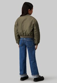 Giubbotto bomber verde oliva con polsini a coste, abbinato a jeans blu larghi con orli arrotolati e due tasche posteriori, indossato con scarpe nere.