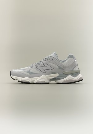Szaro-biały sportowy sneaker z siateczkową i syntetyczną cholewką, sznurowany, z amortyzowaną podeszwą i markowym logo po bokach na neutralnym tle.