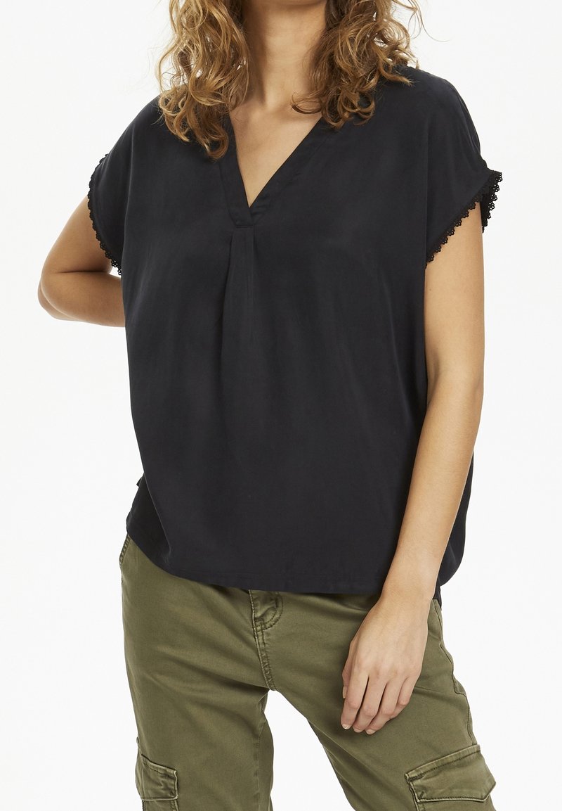 Nainen kiharatukkaisena, pukeutunut löysään mustaan V-kauluksiseen puseroon, jonka hihat on koristeltu pitsireunuksilla, ja oliivinvihreisiin cargo-housuihin, seisoo käsivarsi lanteilla.